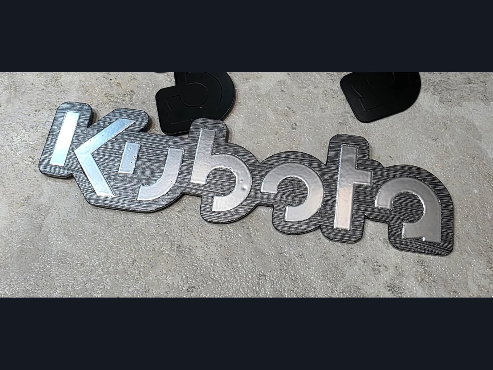 Шильдик с серебристыми буквами Kubota на тёмном фигурном основании и серой рабочей поверхности.