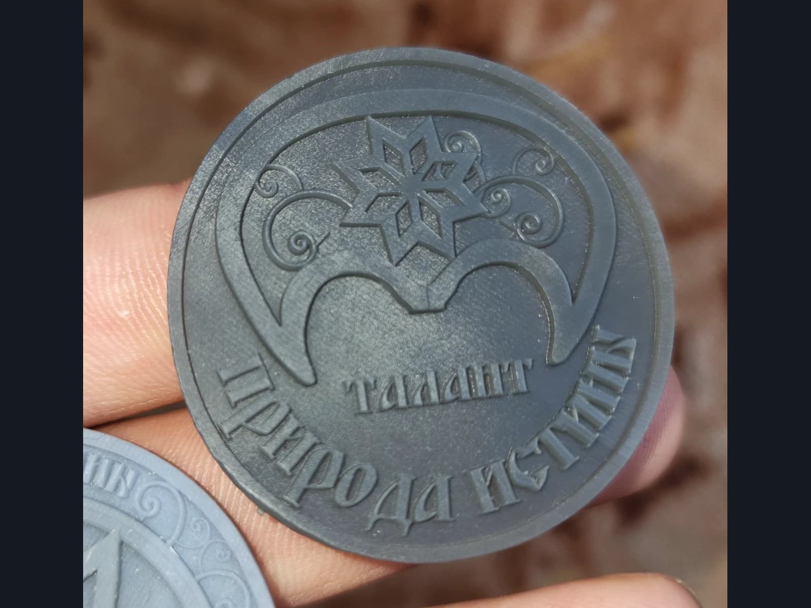 Серый рельефный жетон «Талант» с орнаментом и надписью «Природа истины» в руке.