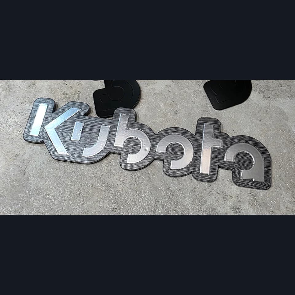 Шильдик с серебристыми буквами Kubota на тёмном фигурном основании и серой рабочей поверхности.